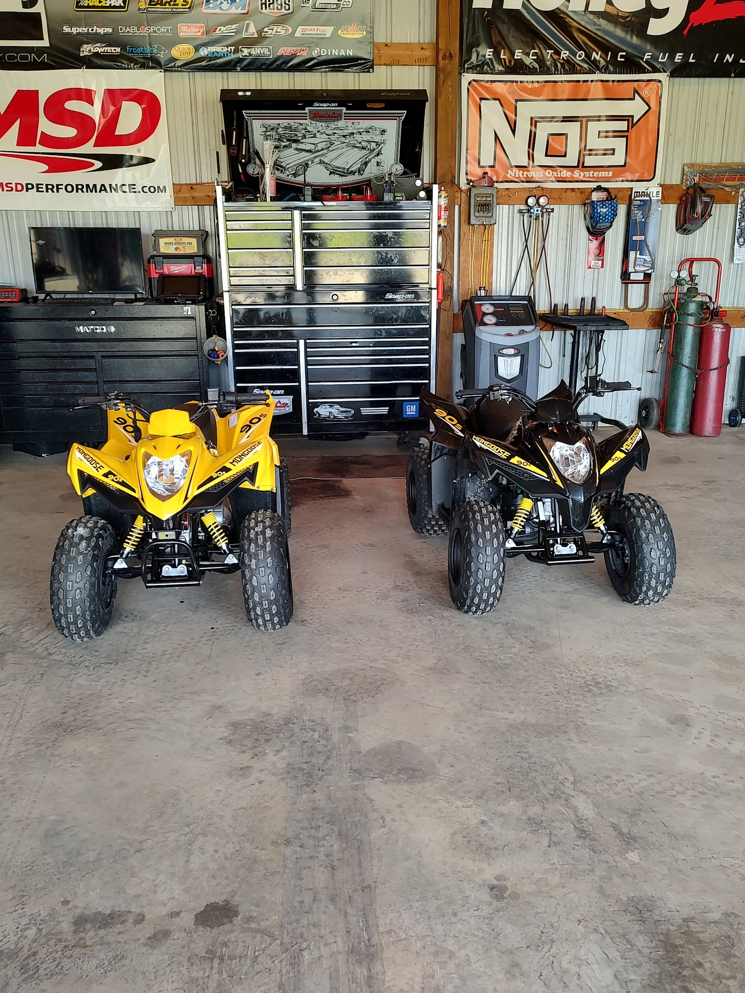 Two Mongoose ATVs, yellow and black, inside the shop with Snap-on toolbox and performance banners