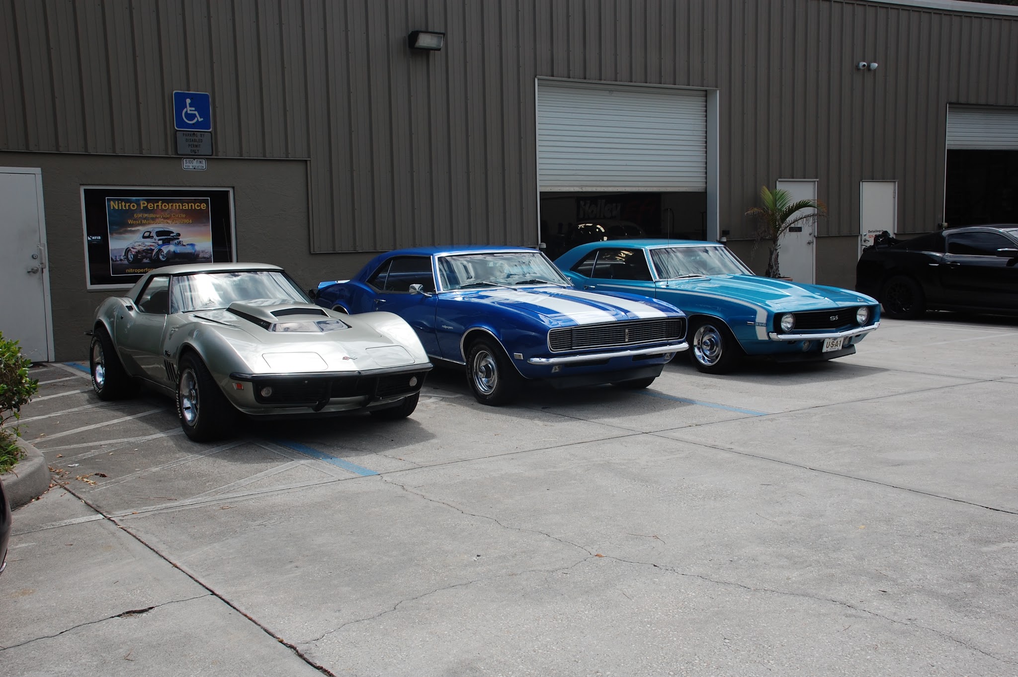 Three classic cars outside the shop: Corvette Stingray, Camaro with racing stripes, and Camaro SS